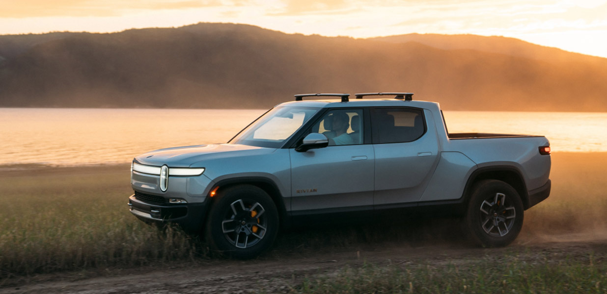 A Rivian electric pickup trick drives along a dirt road next to a lake at sunset