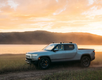 A Rivian electric pickup trick drives along a dirt road next to a lake at sunset