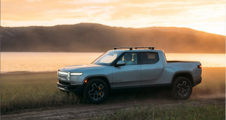 A Rivian electric pickup trick drives along a dirt road next to a lake at sunset