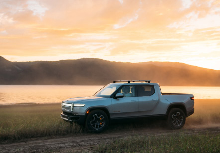 A Rivian electric pickup trick drives along a dirt road next to a lake at sunset