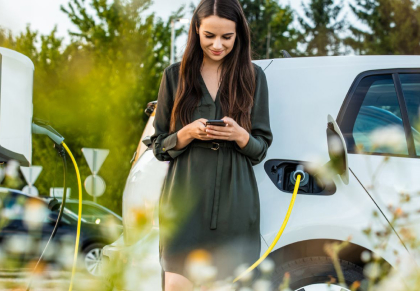 A woman uses her cell phone while her electric vehicle charges behind her