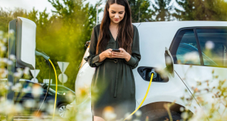 A woman uses her cell phone while her electric vehicle charges behind her