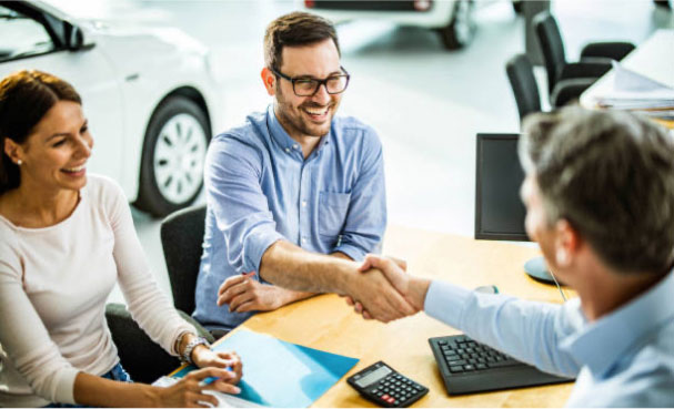 A man sitting next to a woman and across from an employee at an electric vehicle dealership, shaking the employee's hand