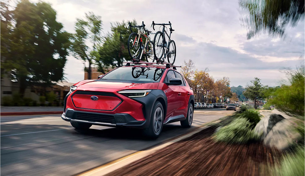 A Subaru electric vehicle with two bikes secured to the roof speeds down a wooded road